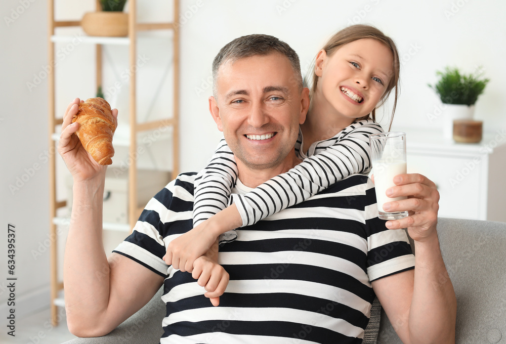 Little girl and her father with croissant drinking milk in living room