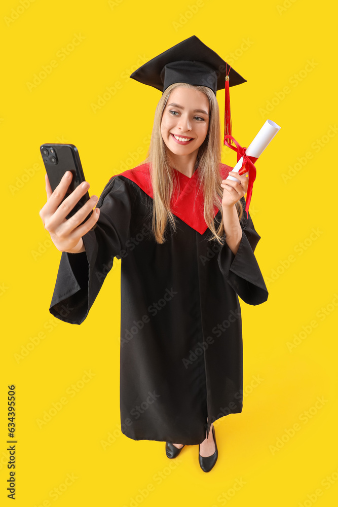 Female graduate student with diploma taking selfie on yellow background