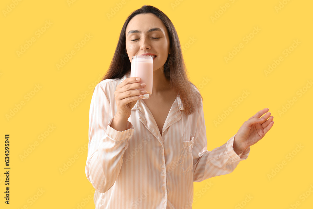 Young woman in pajamas drinking tasty yoghurt on yellow background