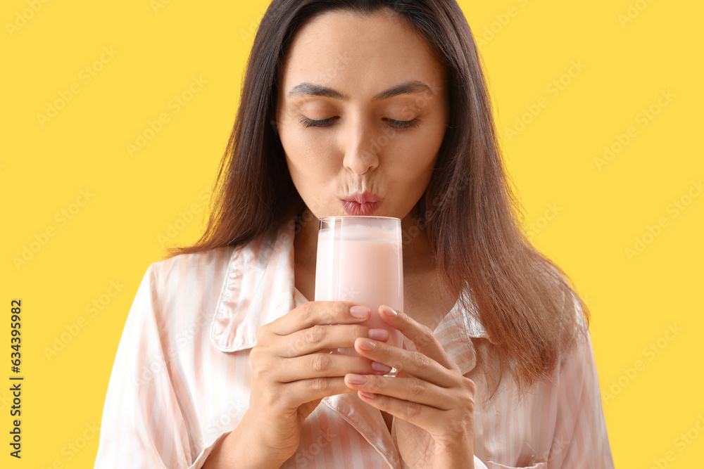 Young woman in pajamas drinking tasty yoghurt on yellow background, closeup