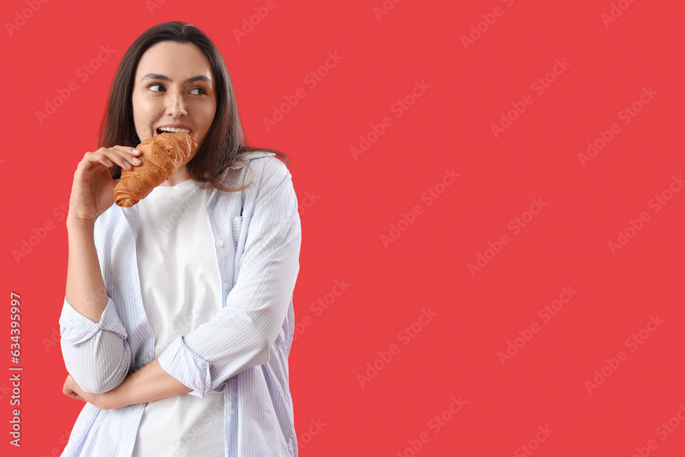 Young woman eating tasty croissant on red background