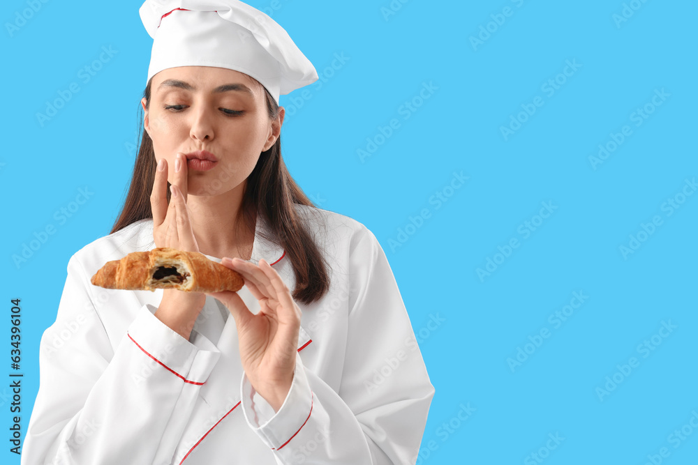 Female chef eating tasty croissant on blue background