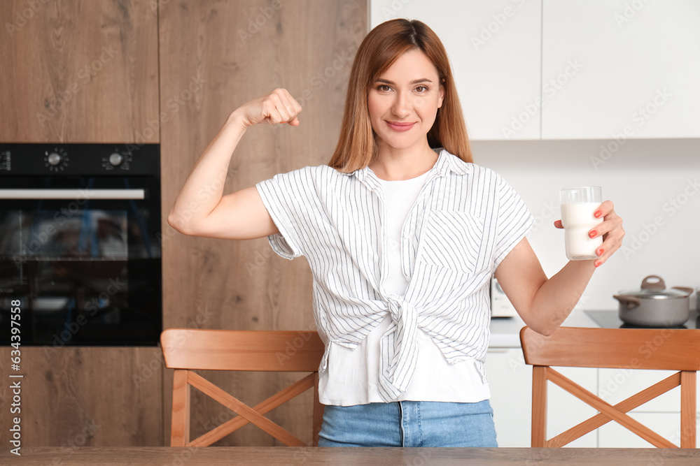 Young woman with glass of milk showing muscles in kitchen