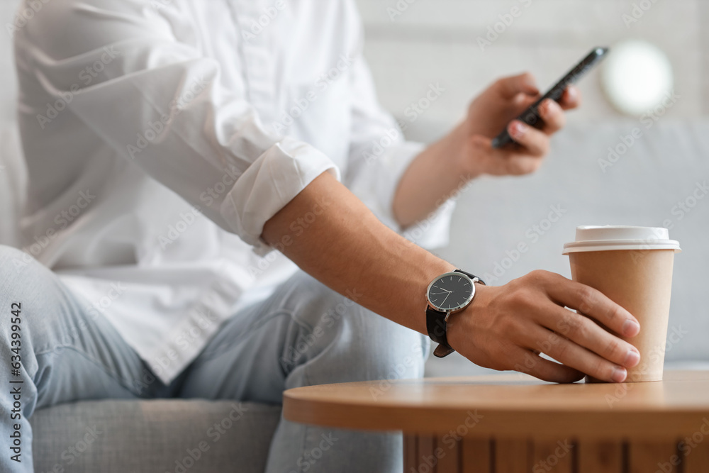 Young man with wristwatch and mobile phone taking cup of coffee at home, closeup