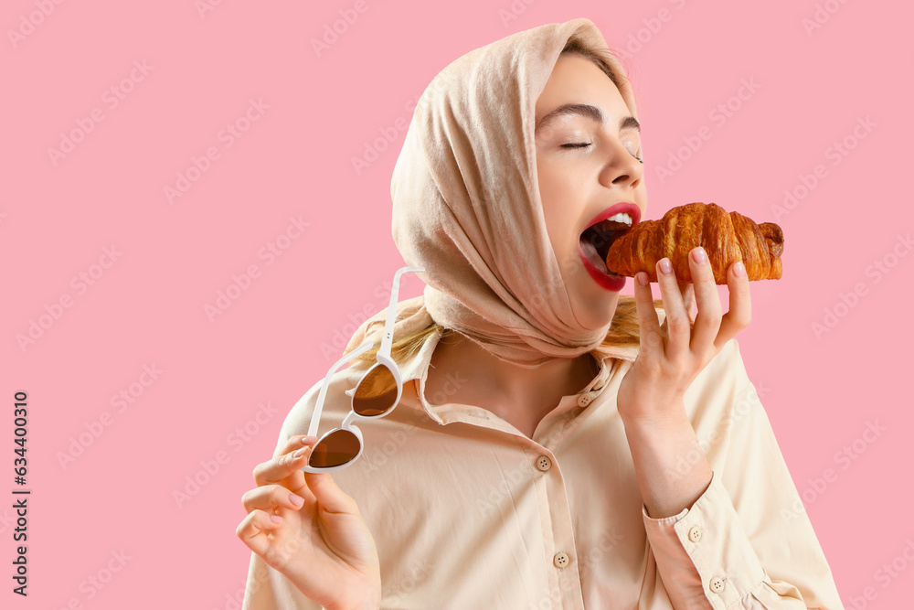 Beautiful stylish young woman eating tasty croissant on pink background