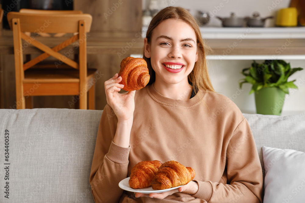 Beautiful young woman sitting on sofa and holding plate with tasty croissants in living room