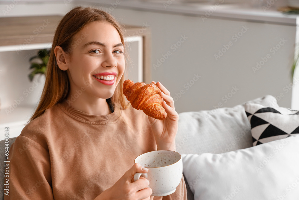 Beautiful happy young woman with tasty croissant and cup of coffee sitting on sofa in living room