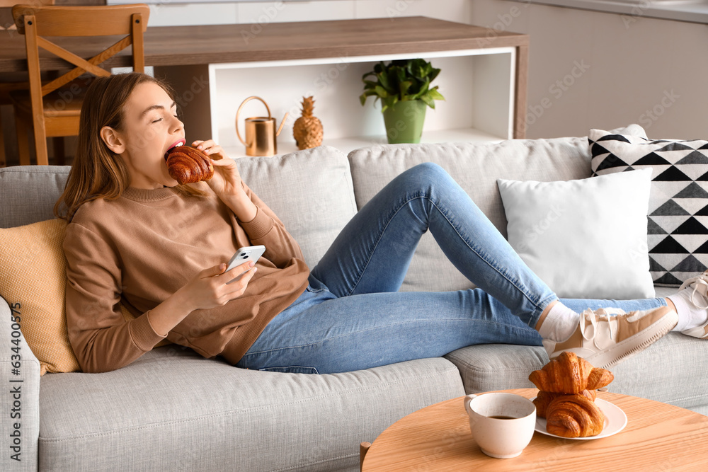 Beautiful young woman eating tasty croissant and using mobile phone on sofa in living room