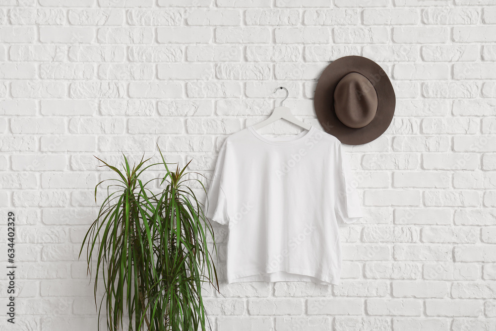 Stylish t-shirt and hat hanging on white brick wall