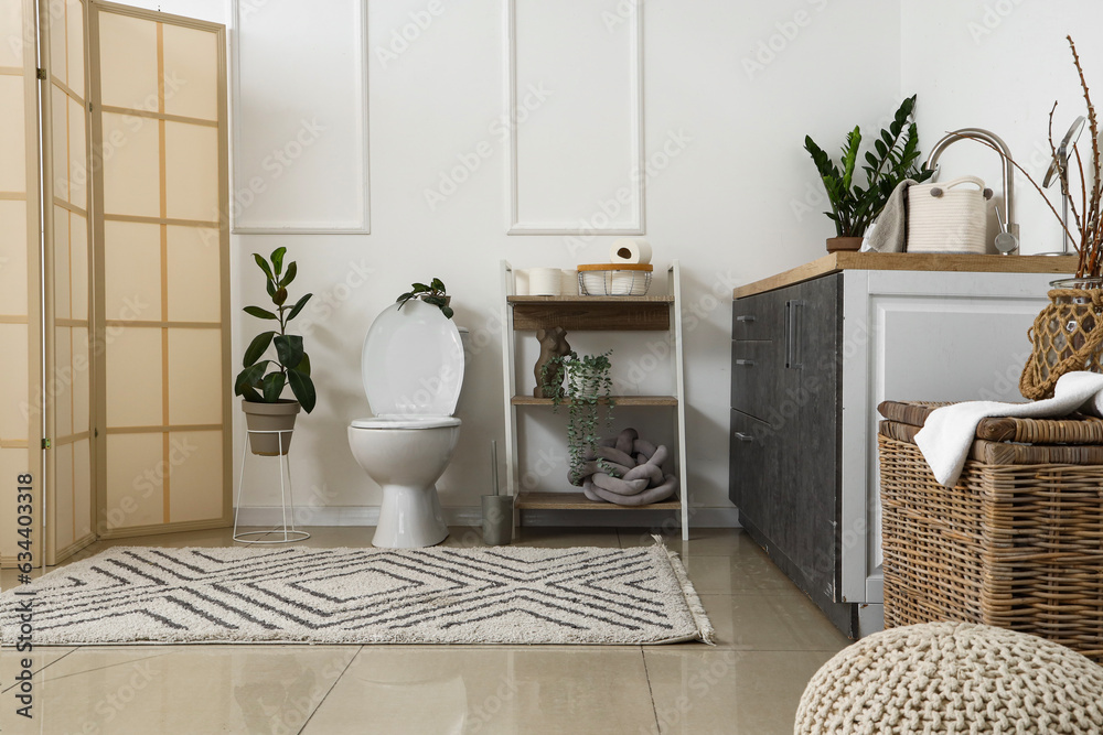 Interior of light restroom with ceramic toilet bowl, shelving unit and houseplants