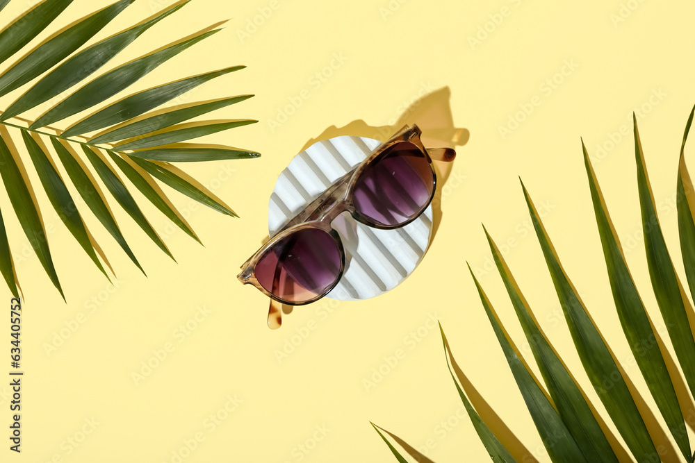 Stand with stylish print sunglasses and palm leaves on pale yellow background