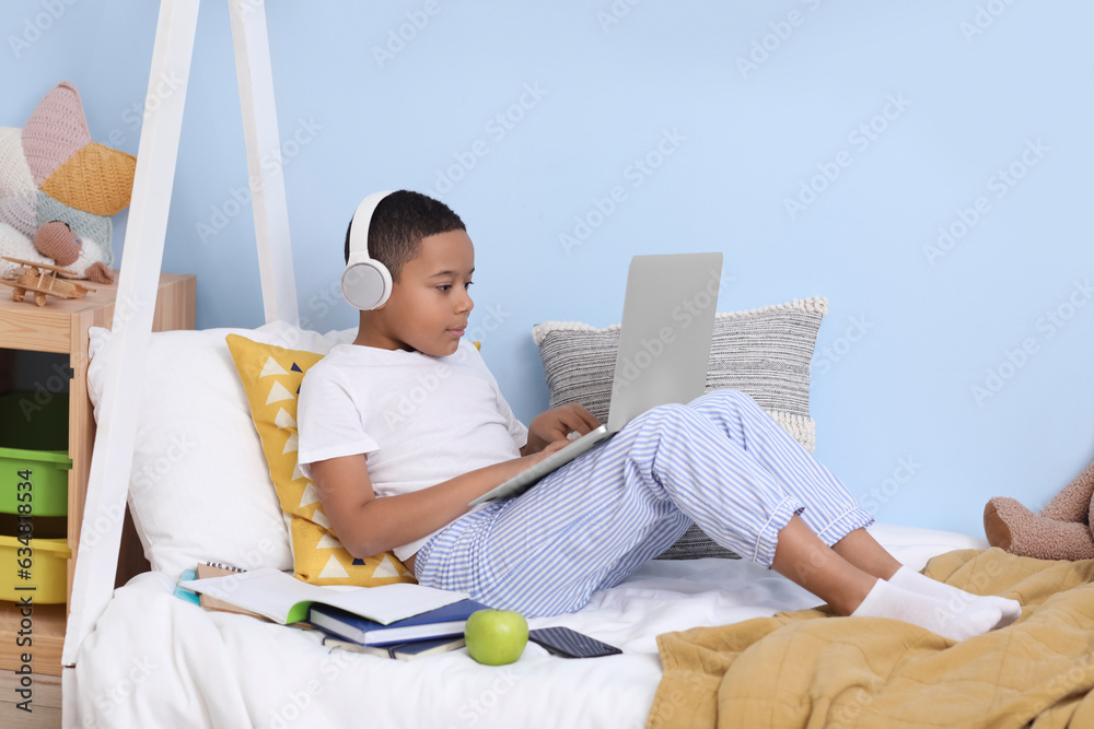 Little African-American boy with laptop studying computer sciences online in bedroom