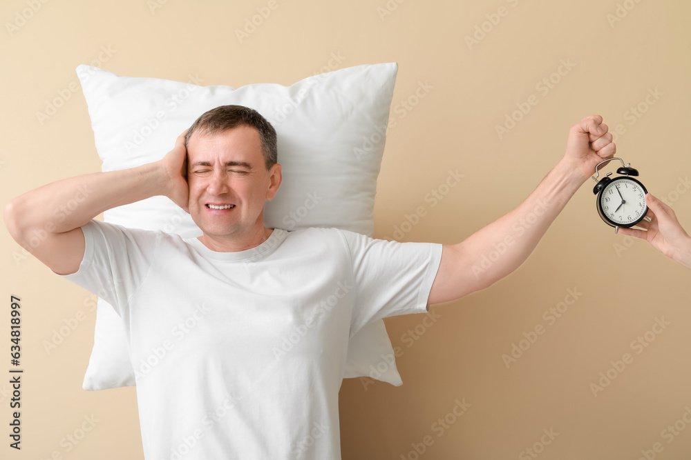 Displeased mature man and female hand with alarm clock on beige background