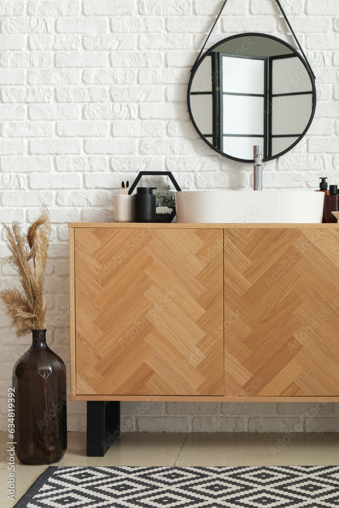 Wooden cabinet with sink bowl and bath accessories near white brick wall