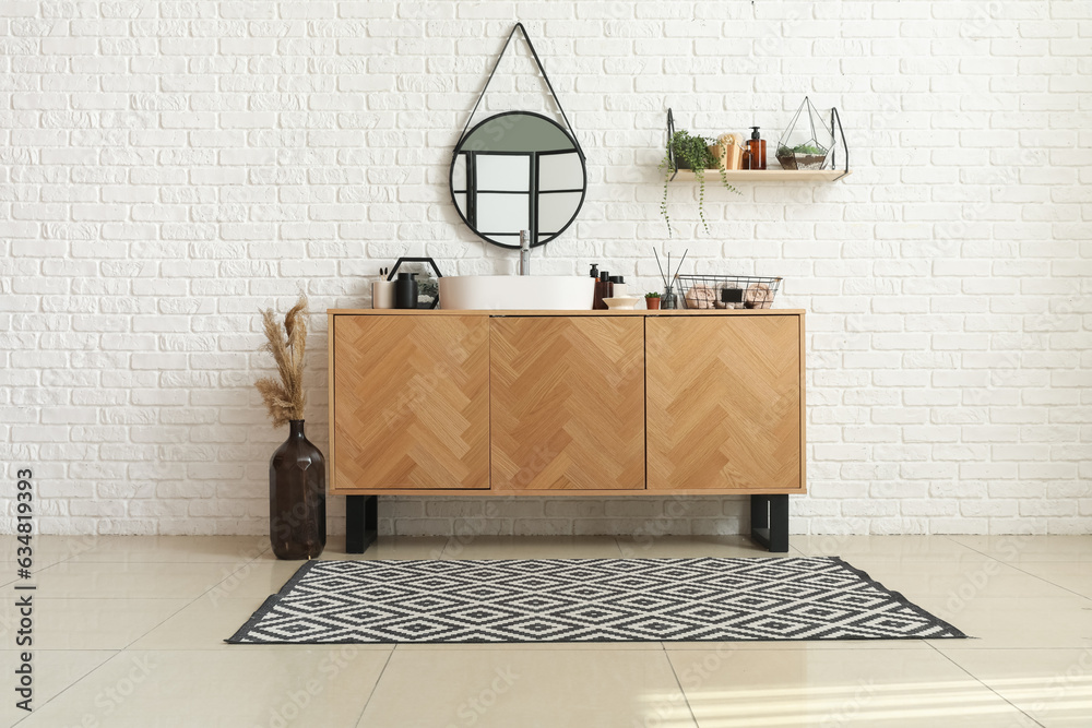 Wooden cabinet with sink bowl and bath accessories near white brick wall