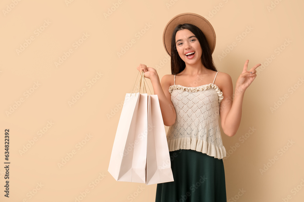 Happy beautiful young woman with shopping bags showing victory gesture on beige background