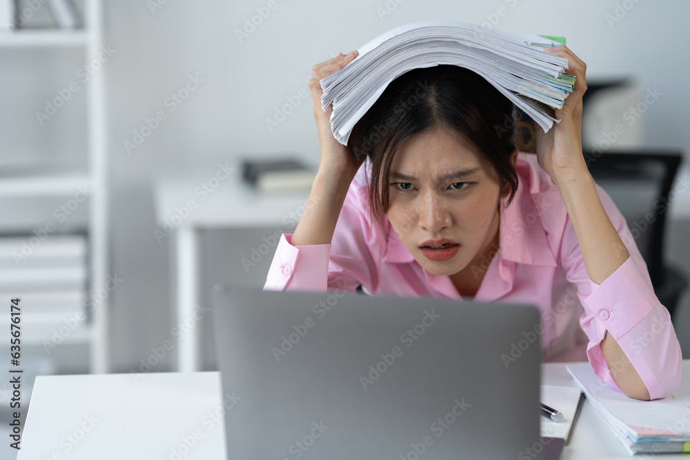 Asian female office worker stressed while working with stacks of documents and laptop. Feeling tired