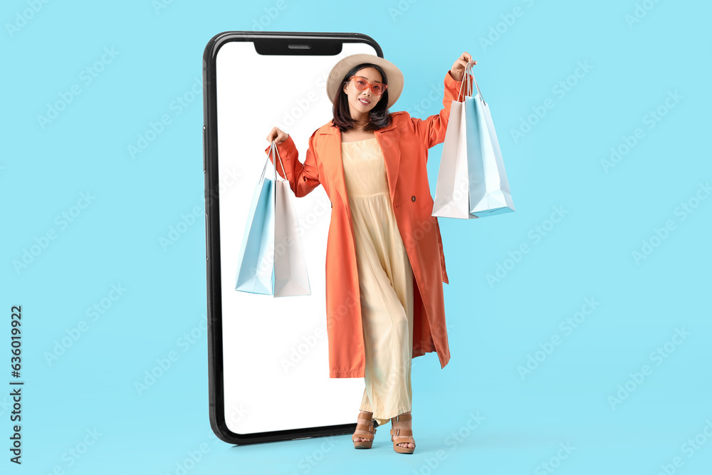 Stylish Asian woman with shopping bags near big mobile phone on light blue background