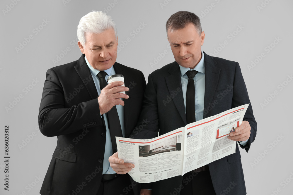 Mature brothers with cup of coffee reading newspaper on grey background