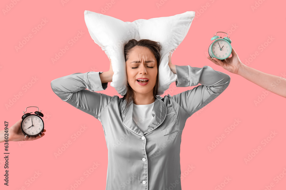 Displeased young woman with pillow and hands holding alarm clocks on pink background