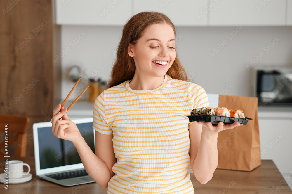 Happy young woman with laptop eating sushi in kitchen