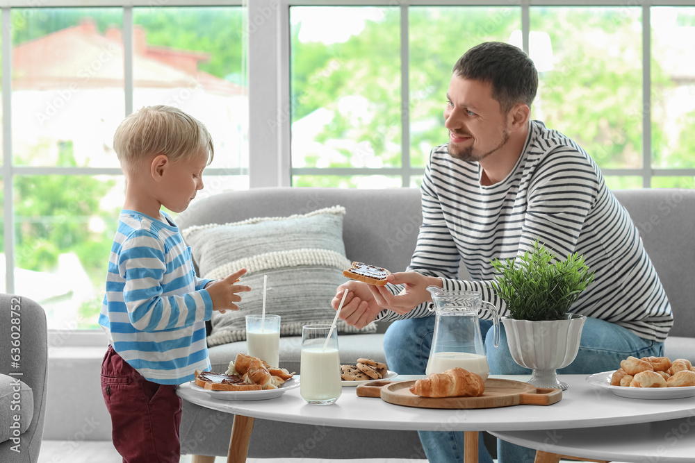 Little boy with his father eating different pastries and drinking milk at table in living room