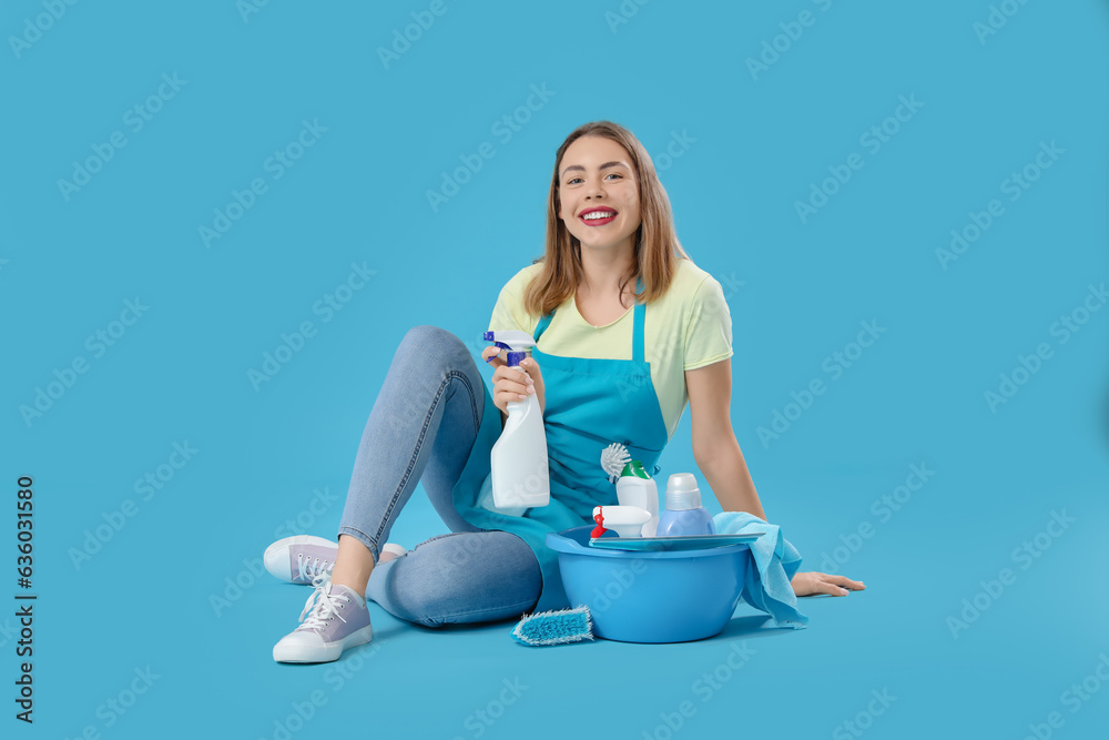 Young woman with cleaning supplies sitting on blue background