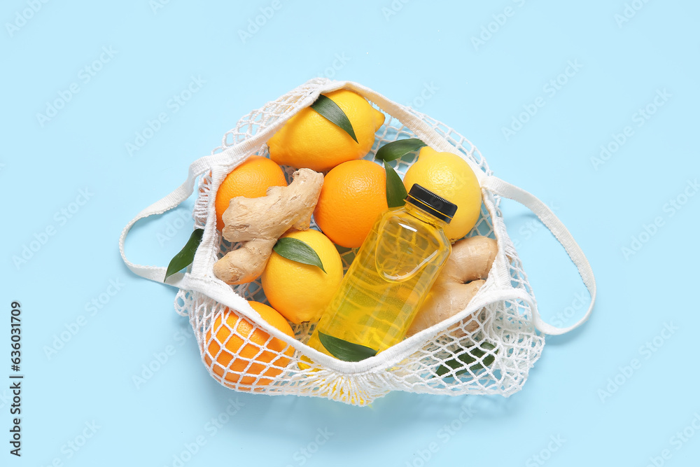 Mesh bag with fresh lemons, oranges, ginger and bottle of juice on blue background