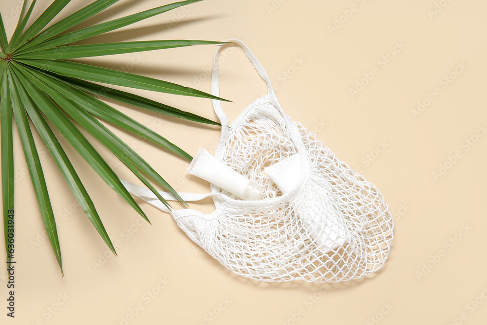 Mesh bag with palm leaf and tubes of cosmetic products on beige background