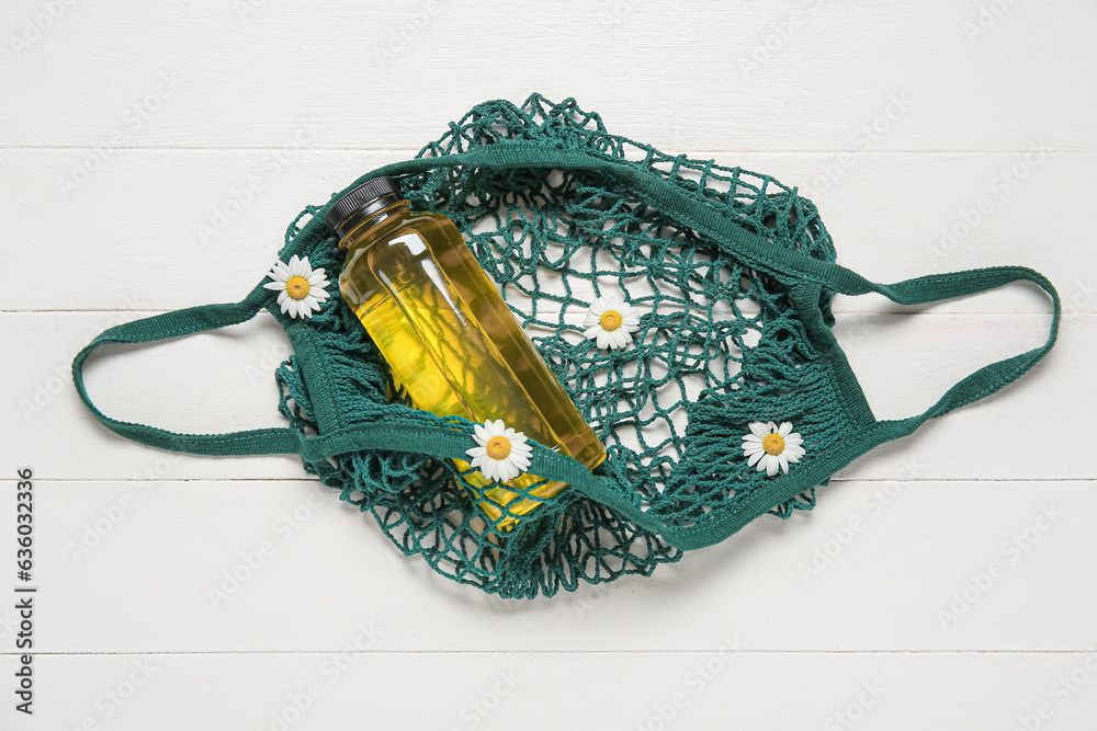 Mesh bag with bottle of juice and beautiful chamomile flowers on light wooden background