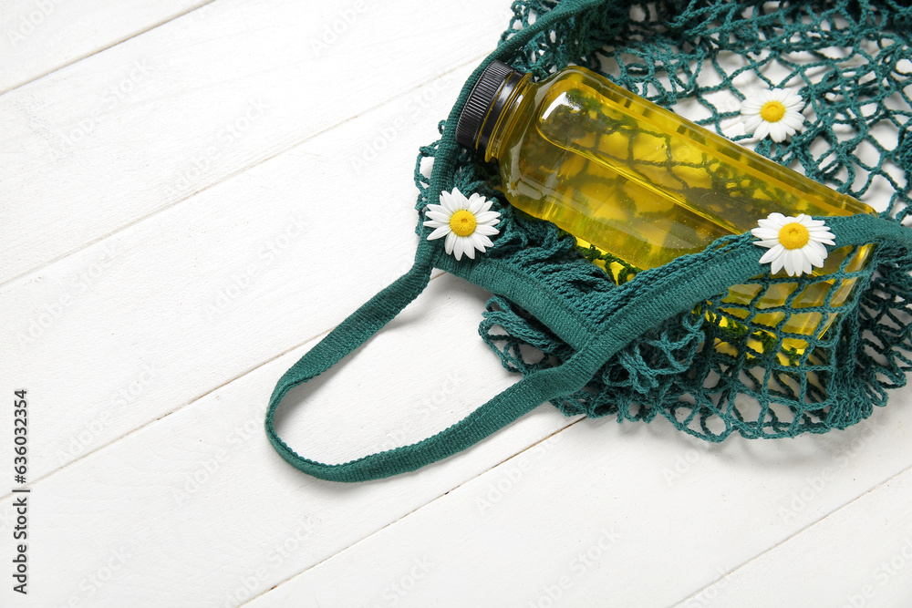 Mesh bag with bottle of juice and beautiful chamomile flowers on light wooden background, closeup