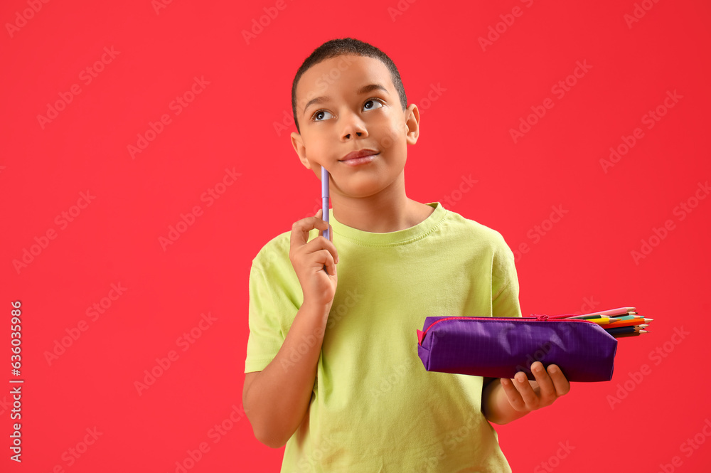 Thoughtful little African-American boy with pen and pencil case on red background