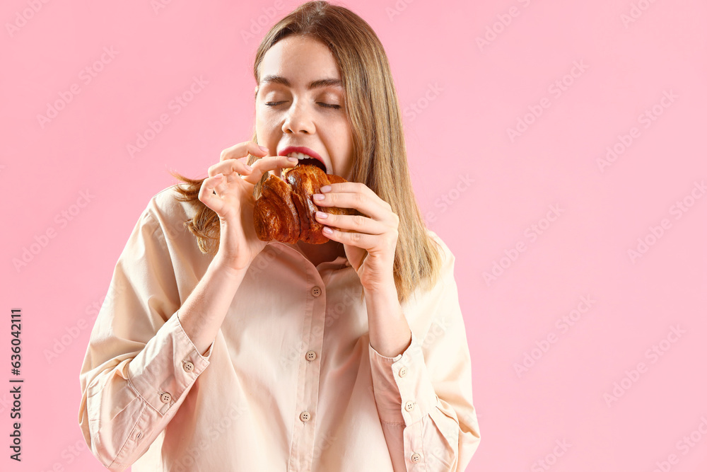 Beautiful young woman eating tasty croissant on pink background