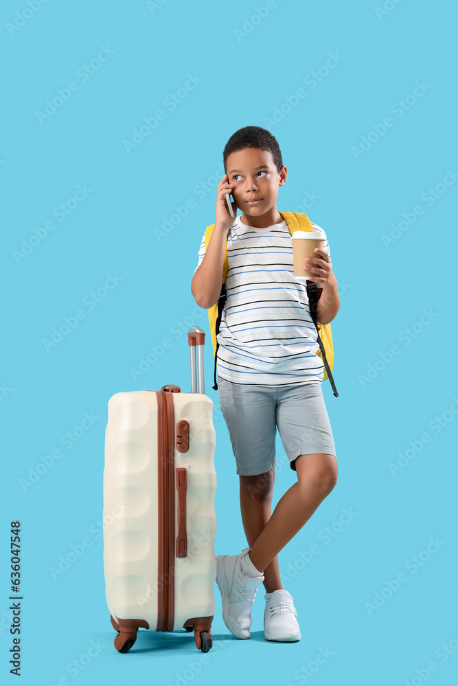 Little African-American boy with suitcase and cup of coffee talking by phone on blue background