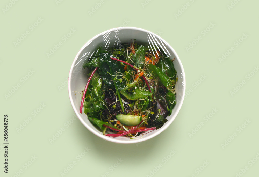 Bowl of fresh salad covered with plastic food wrap on green background