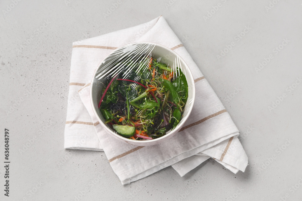 Bowl of fresh salad covered with plastic food wrap on grey background