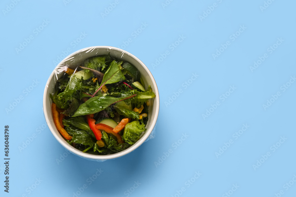 Bowl of fresh salad covered with plastic food wrap on blue background