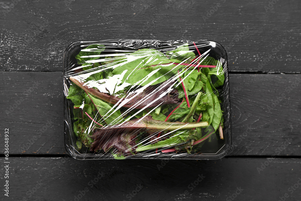 Fresh salad leaves covered with plastic food wrap on black wooden background