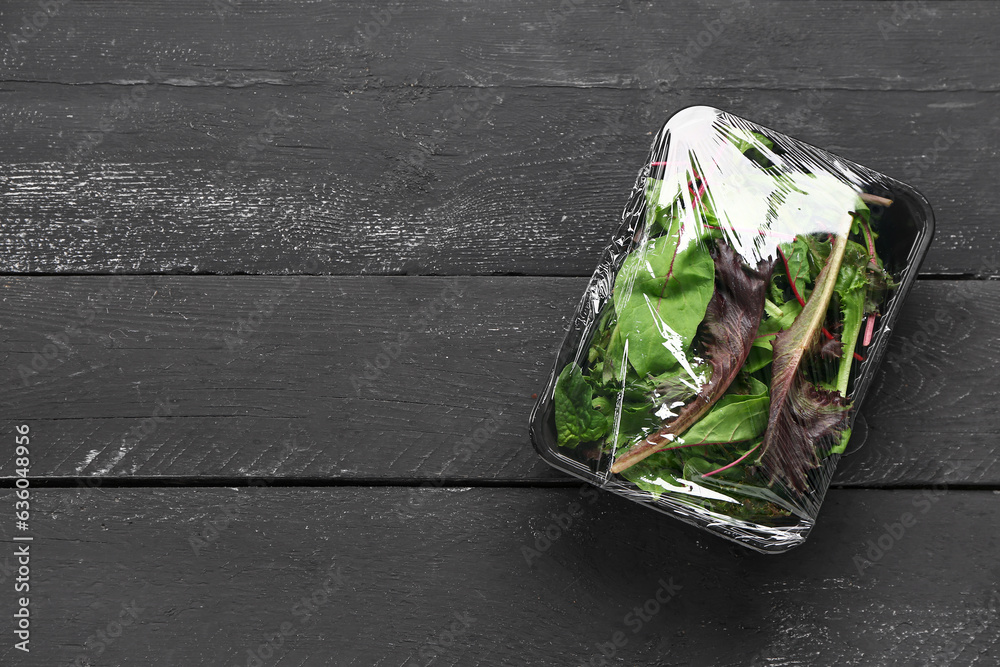 Fresh salad leaves covered with plastic food wrap on black wooden background