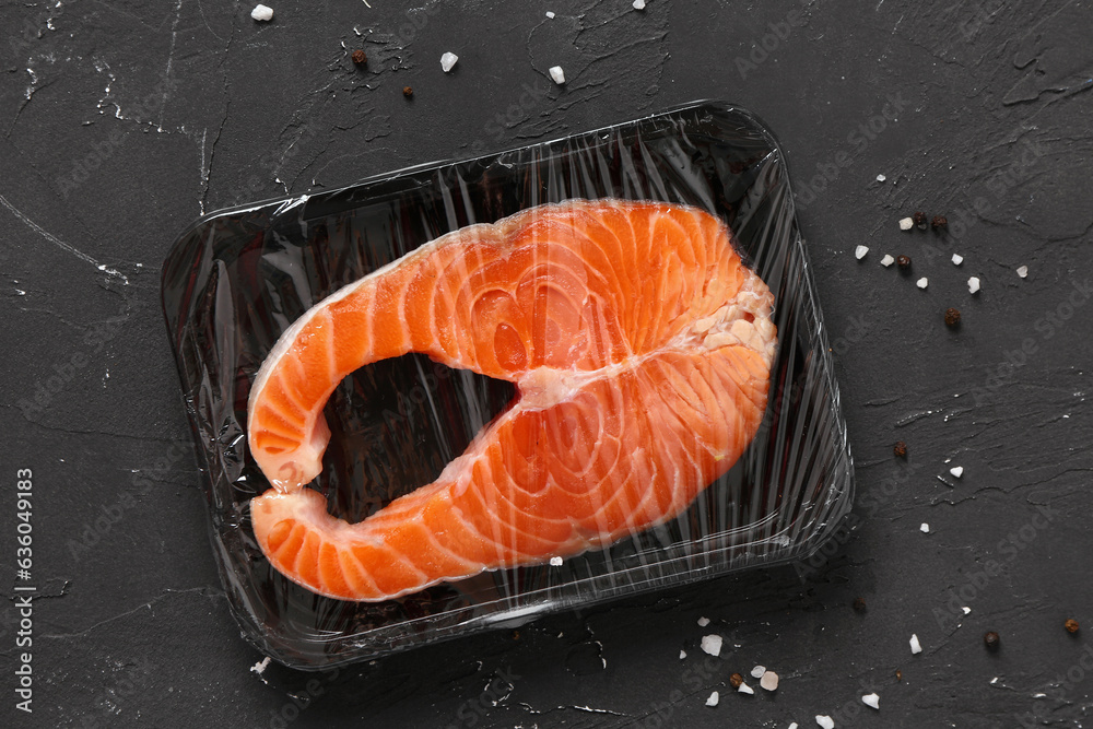 Fresh salmon steak in plastic food wrap and spices on black background