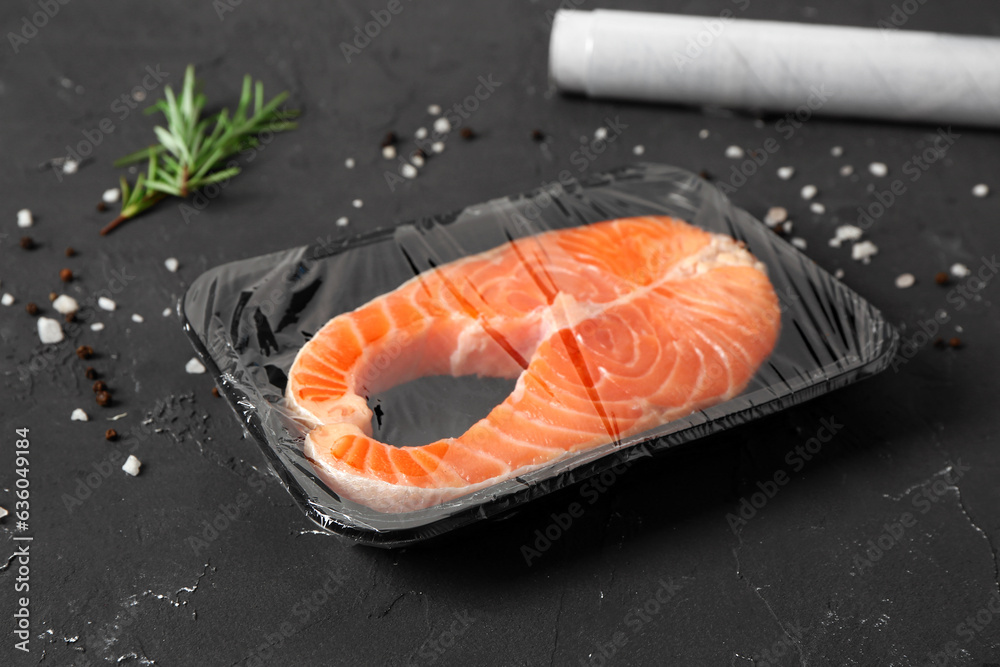 Fresh salmon steak in plastic food wrap and spices on black background