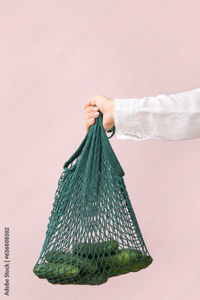 Female hand holding mesh bag with fresh cucumbers on beige background