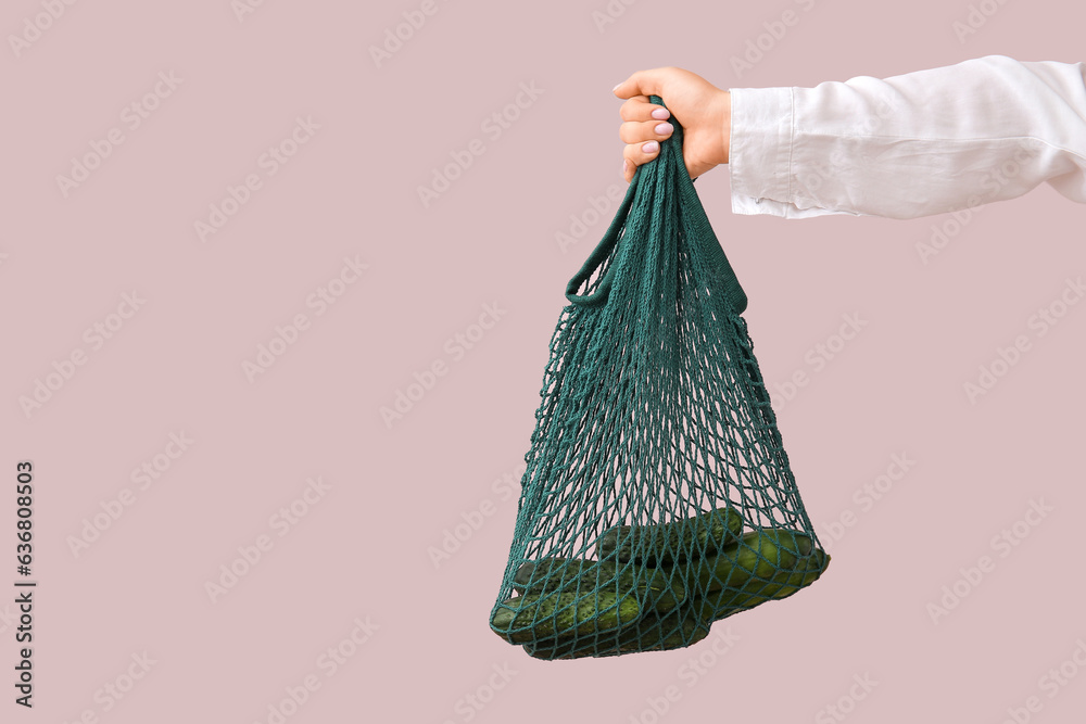 Female hand holding mesh bag with fresh cucumbers on beige background