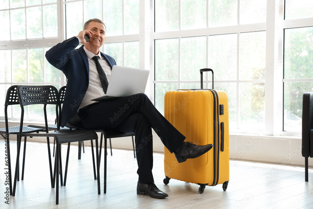 Mature businessman with laptop talking by mobile phone in hall of airport