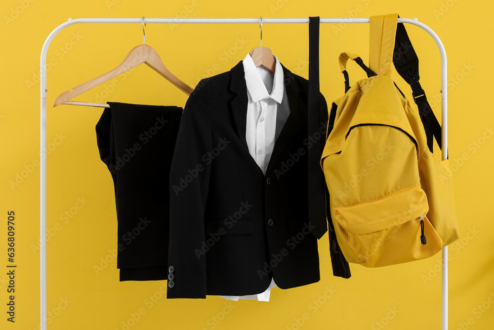 Rack with stylish school uniform and backpack on yellow background