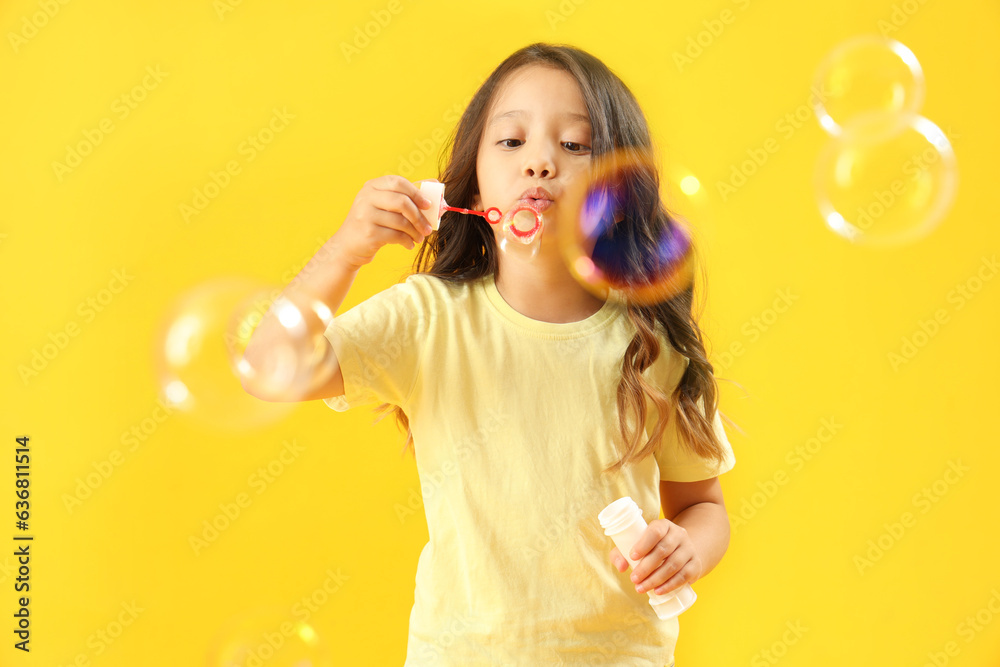 Cute little girl blowing soap bubbles on yellow background