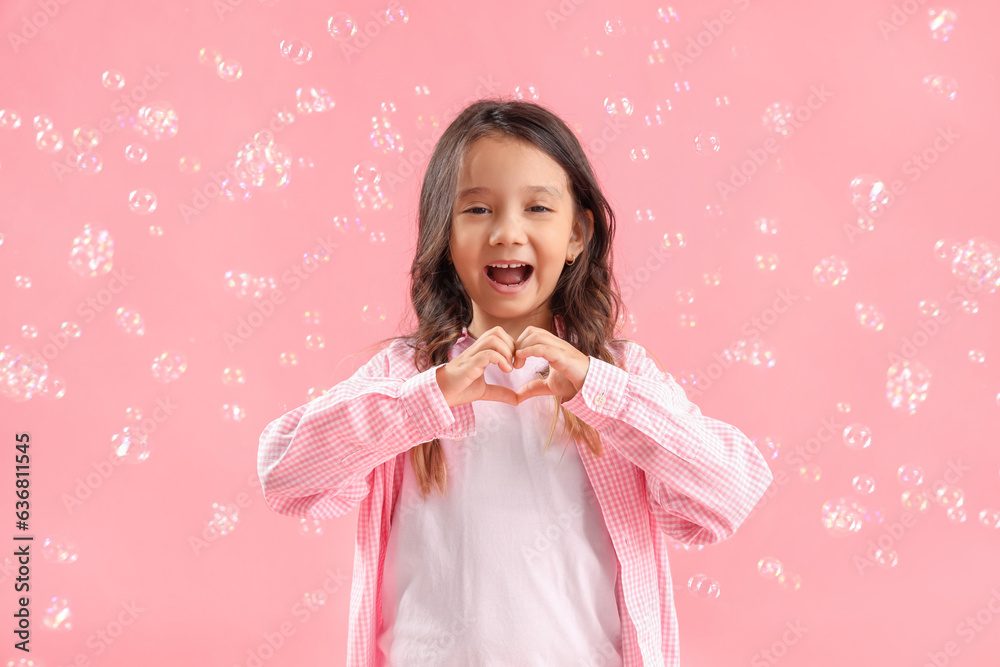 Cute little girl making heart with her hands and soap bubbles on pink background