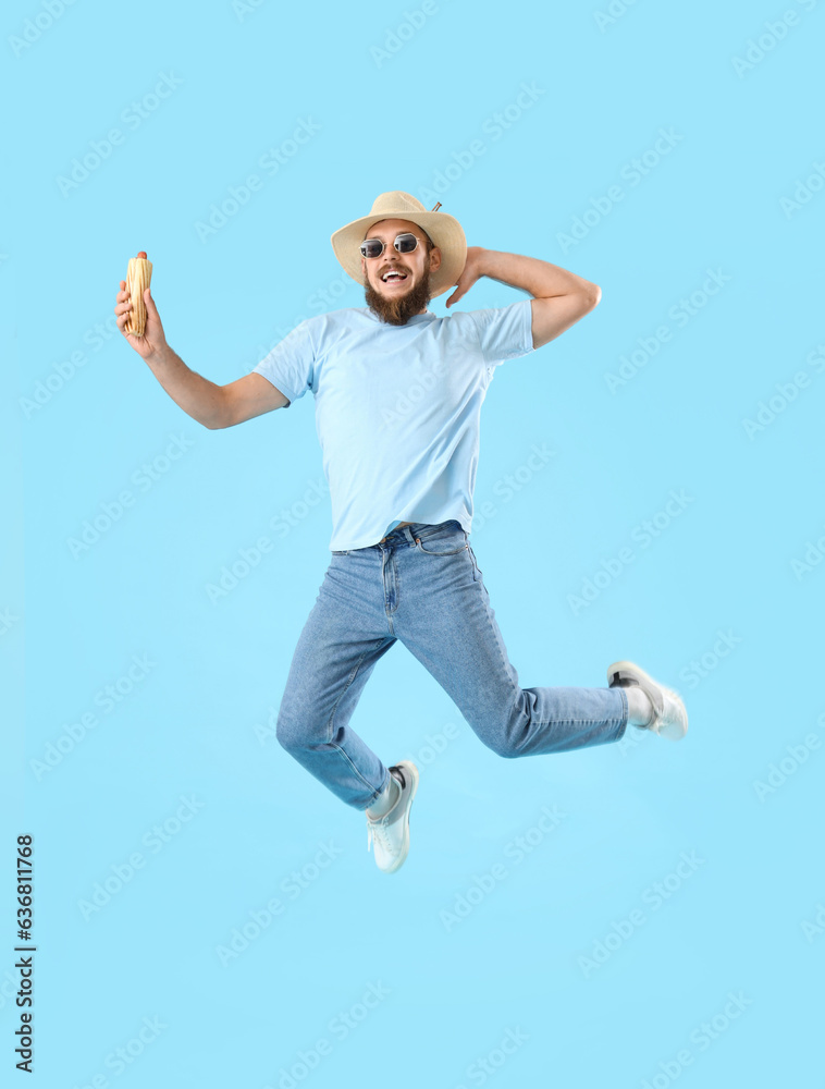 Handsome young man jumping with tasty hot dog on blue background