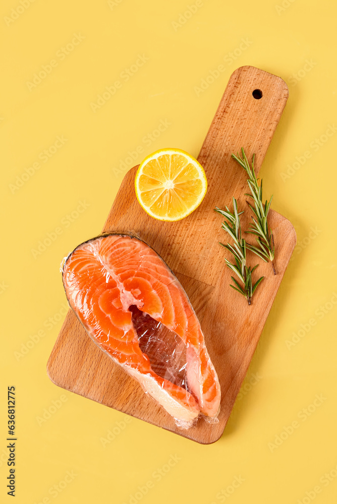 Wooden board of fresh salmon steak in plastic food wrap, lemon and rosemary on yellow background