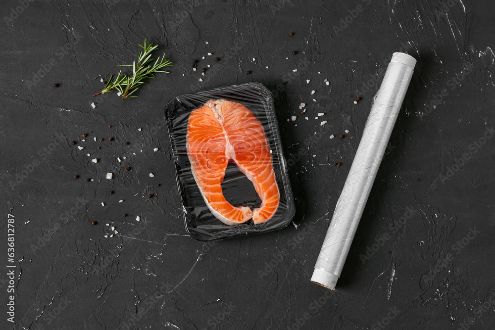 Fresh salmon steak in plastic food wrap and spices on black background